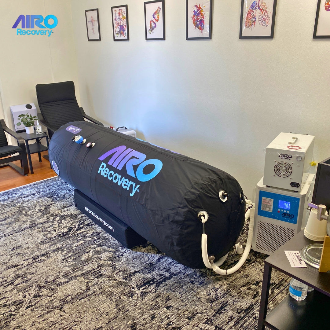Man smiling and standing barefoot next to a hyperbaric oxygen chamber with the AIRO Recovery logo displayed on it
