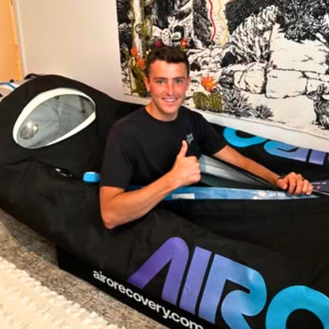 Man smiling and giving a thumbs-up while sitting inside a black and blue hyperbaric chamber labeled 'airorecovery.com' in a cozy indoor setting